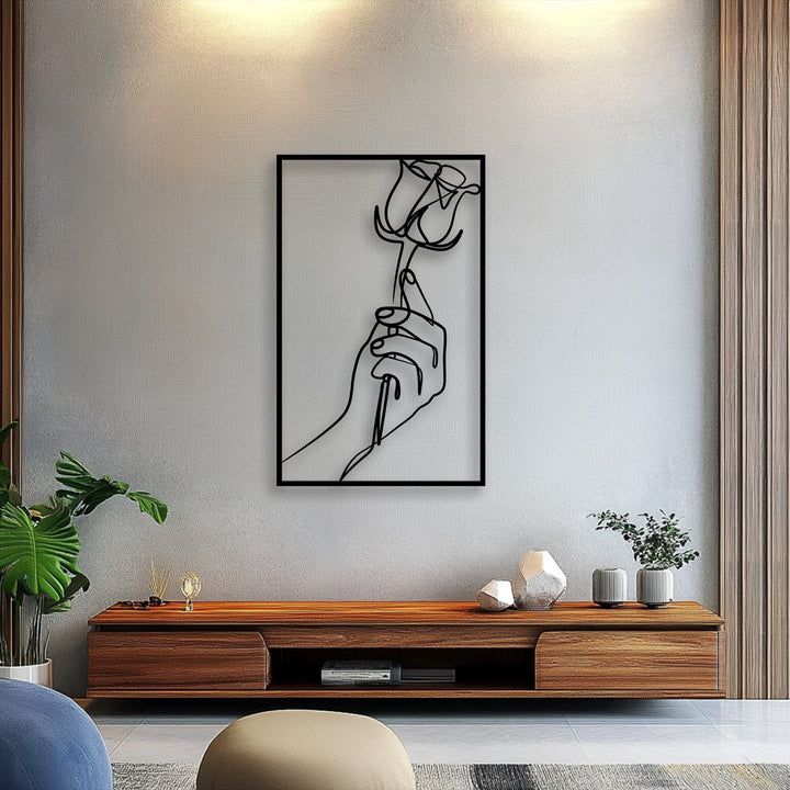 The realistic detail of the elegant line art metal art for wall, portraying a hand holding a rose, is vividly highlighted on the textured wall.