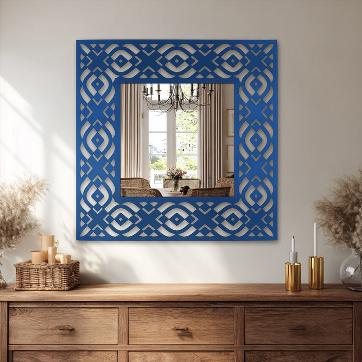 Reflected in the square bathroom mirror with a geometric tribal frame is a warmly lit dining area with a table, chairs, chandelier, and window.