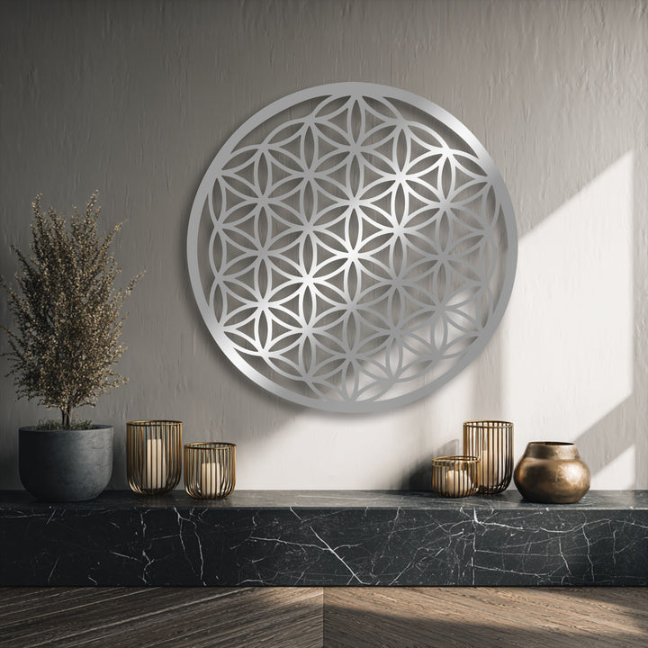 The decorative metal art piece showcases striking color contrast with its luminous silver Flower of Life design against a muted gray wall and deep-toned shelf items.