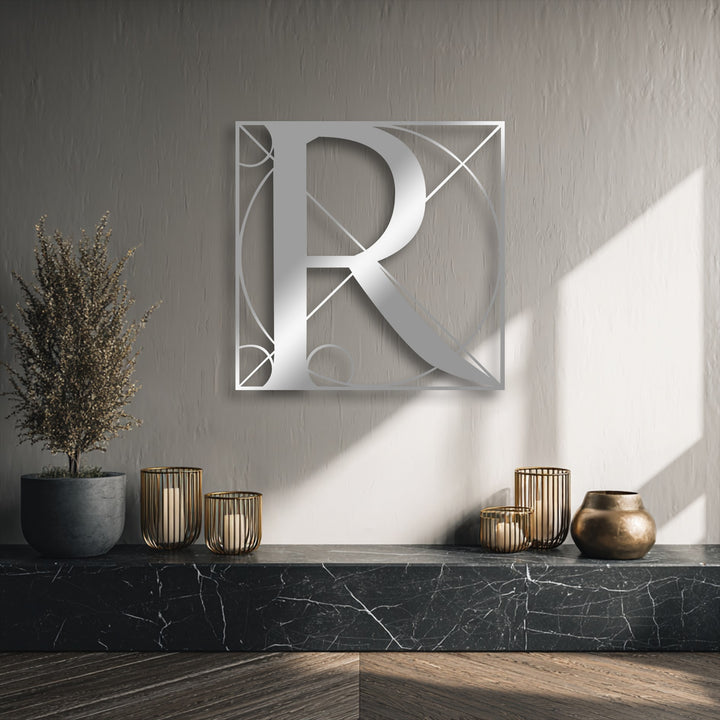 A vertically oriented, square-shaped piece of metal art for wall features a silver geometric letter R design, casting shadows on the textured wall.