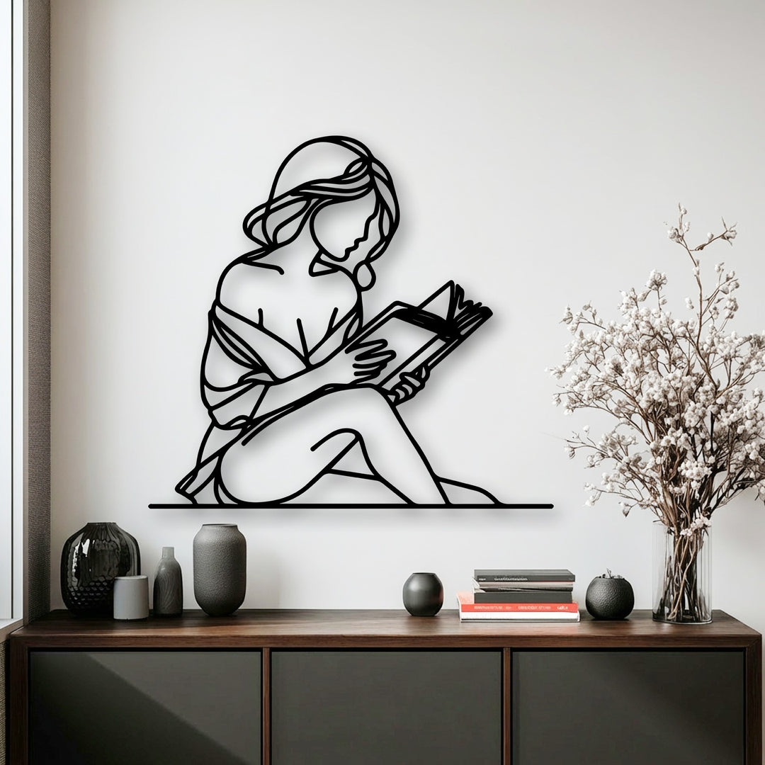A horizontally oriented, gracefully curved silhouette of decorative metal art depicts a minimal woman deeply engrossed in reading a book.