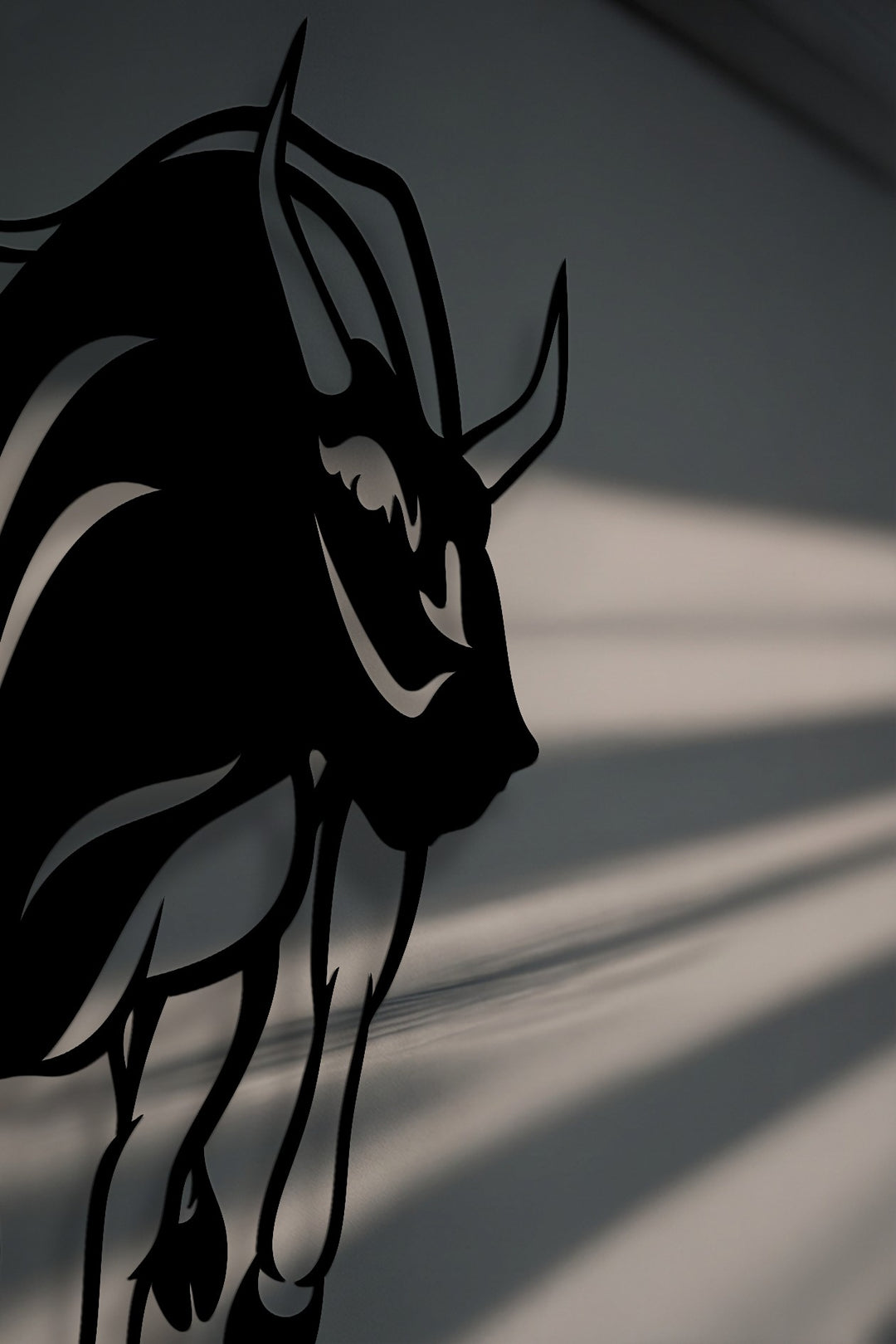 A striking black silhouette of a powerful charging bull, designed as decorative metal art, against a subtly textured grey background with sunlit stripes.