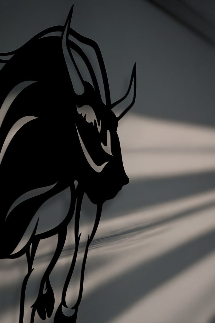 A striking black silhouette of a powerful charging bull, designed as decorative metal art, against a subtly textured grey background with sunlit stripes.