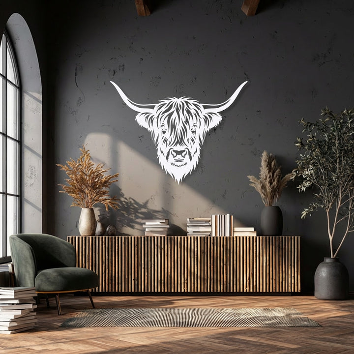 A stack of white books and a dark green armchair rest on the wooden floor, complementing the majestic Highland bull head decorative metal art on the gray wall.