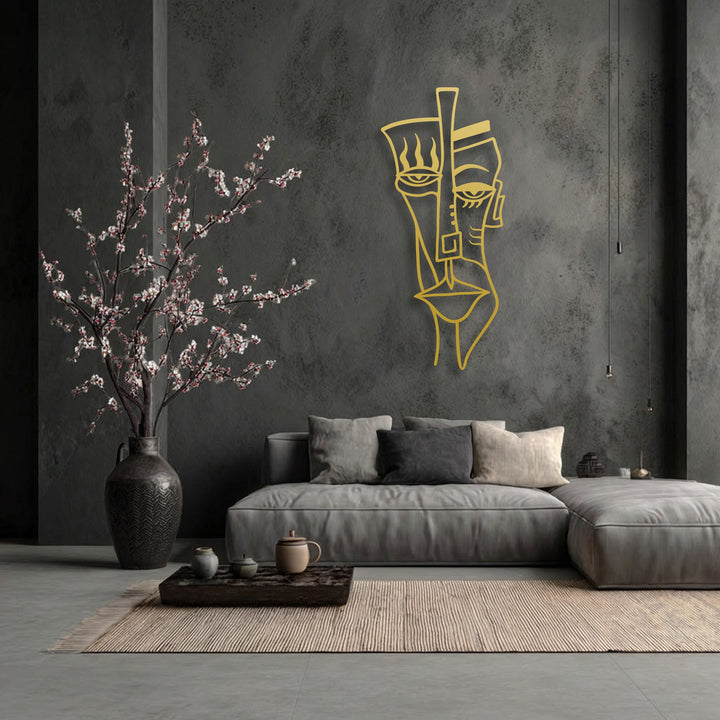 The sharp edges and bold borders of the golden abstract tribal face metal art for wall stand out against the textured dark gray wall.