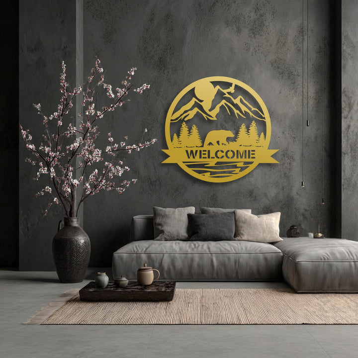 Smooth, golden decorative metal art welcome sign with a bear and mountain against a coarsely textured gray wall and ribbed woven rug.