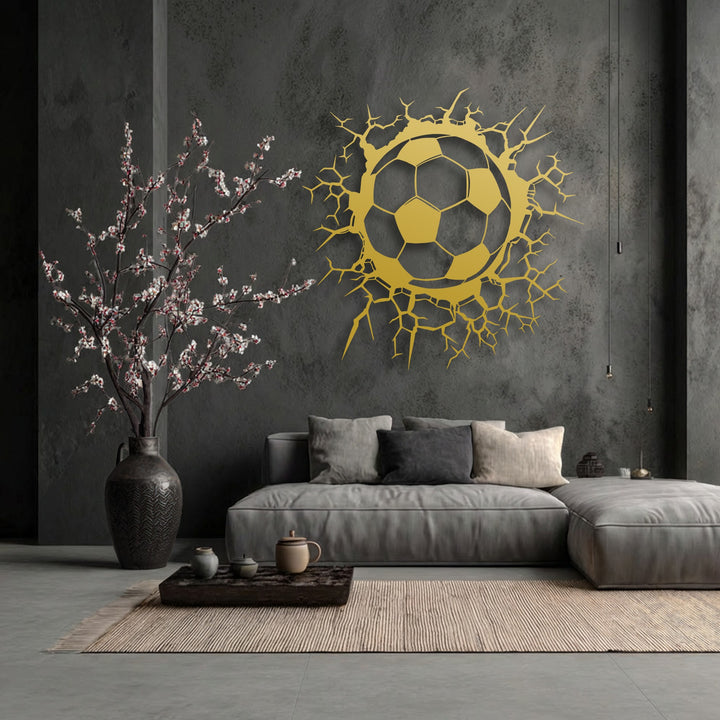 The deep negative space of the textured dark wall dramatically frames the golden breaking wall soccer ball decorative metal art.