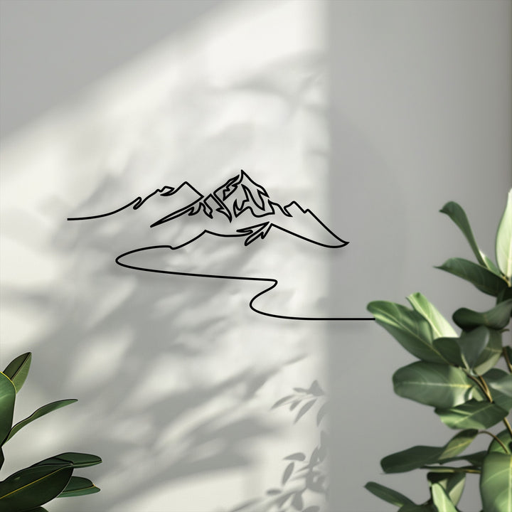 The focal point is a striking black minimalist mountain line decorative metal art piece against a sunlit wall with leaf shadows.