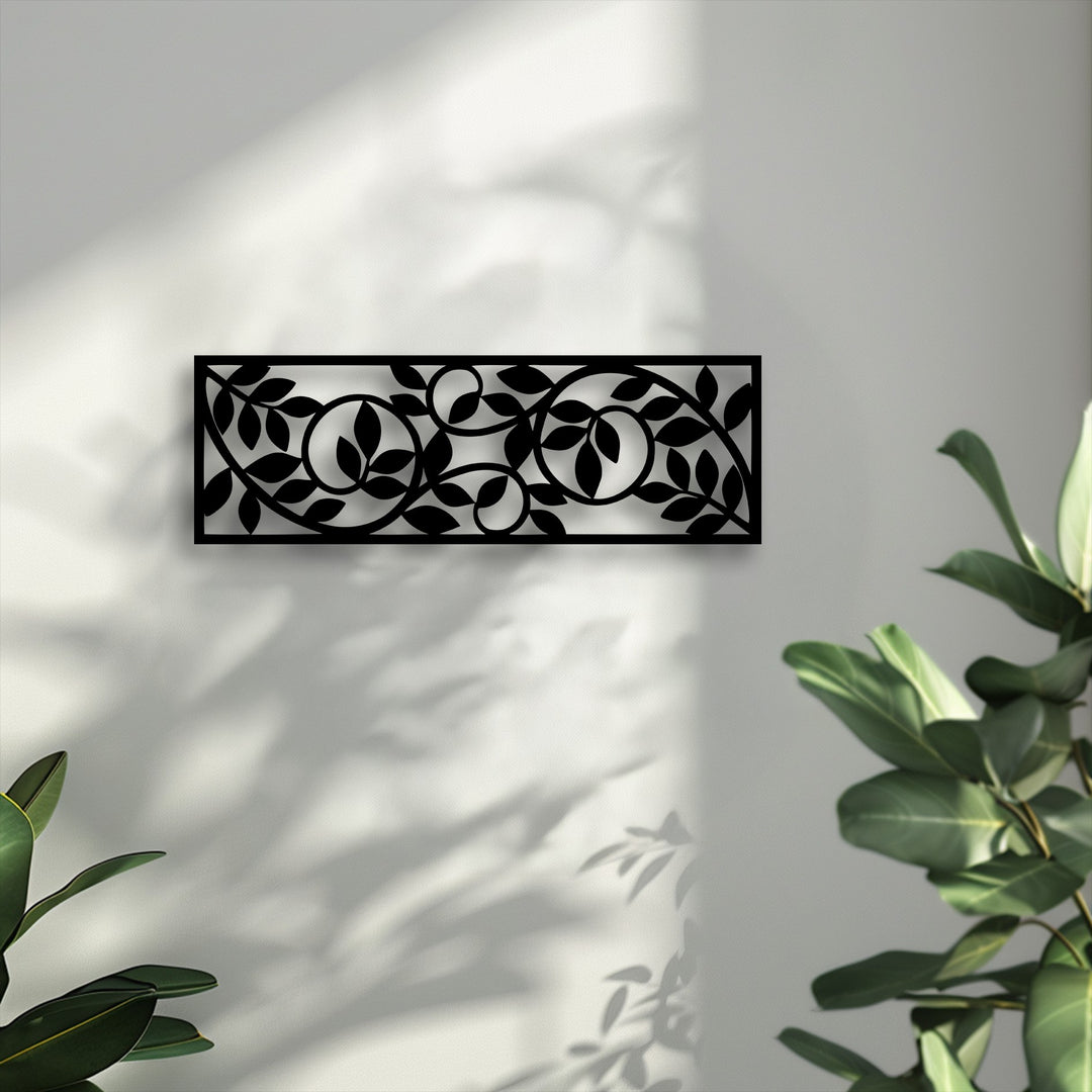 A rectangular black panel of unique metal wall art displays a captivating flowing botanical vine pattern against a sunlit white wall.