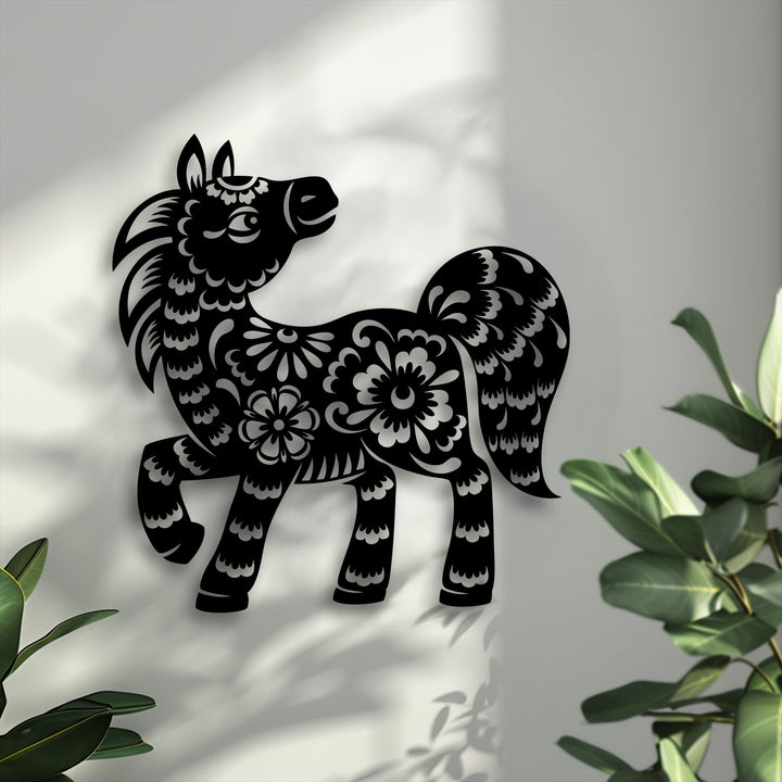 The soft, intricate floral cutouts adorning the black pony metal artwork beautifully contrast the light-filled wall, complemented by blurred green leaves.