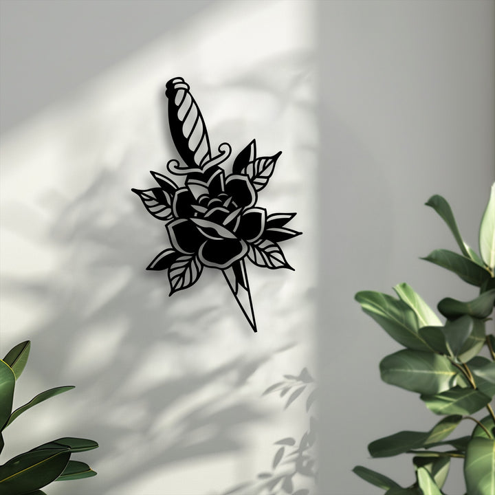 Lush green leaves gently frame a striking black rose and dagger silhouette metal artwork, casting dynamic shadows on a textured white wall.