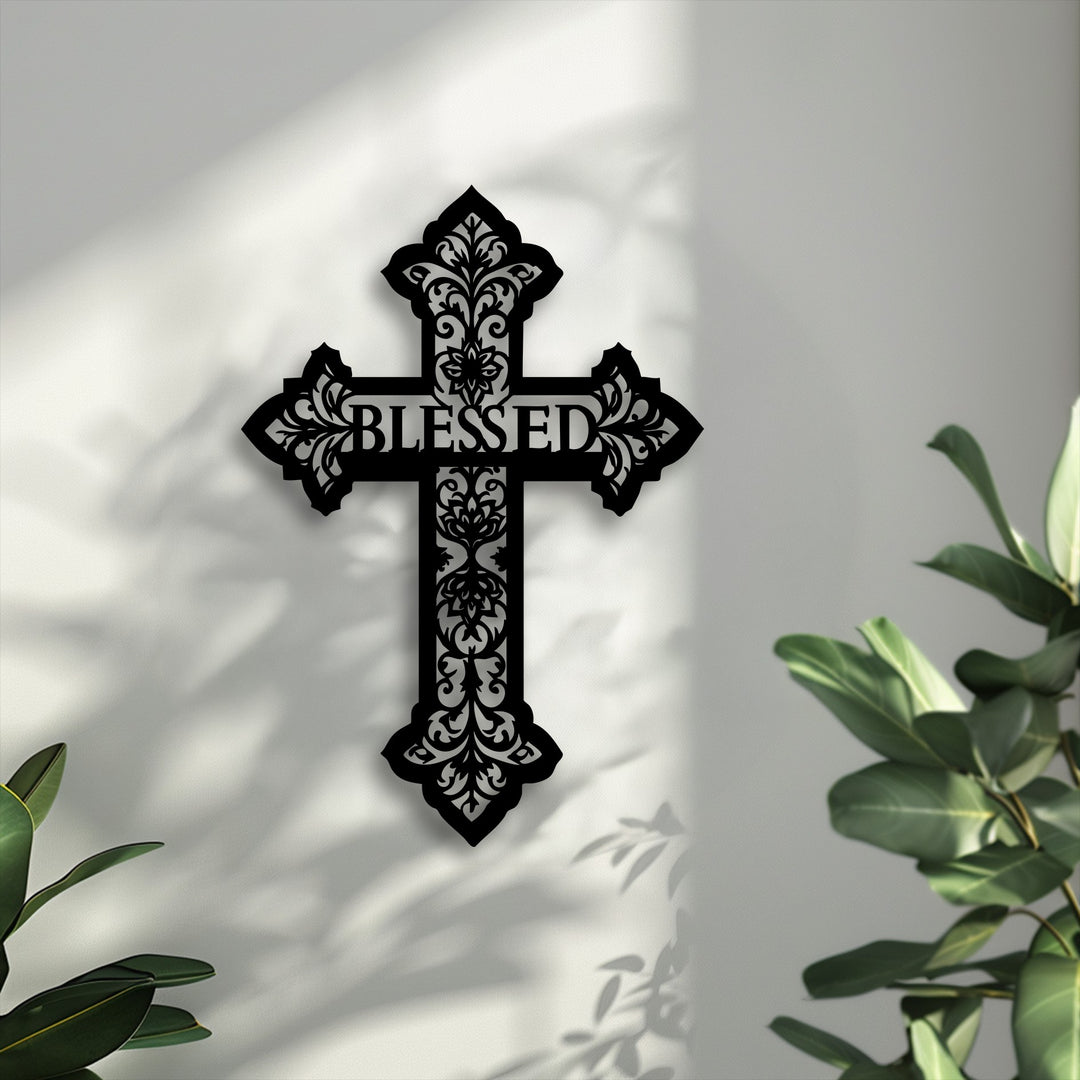 An ornate black Blessed Cross metal artwork featuring intricate scrollwork design casts shadows on a light wall, with green plant leaves nearby.