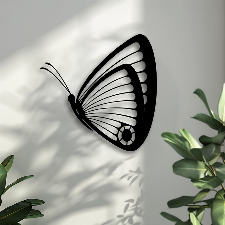 Lush green plant leaves frame the elegant black butterfly metal artwork, casting dynamic shadows on the light gray wall.