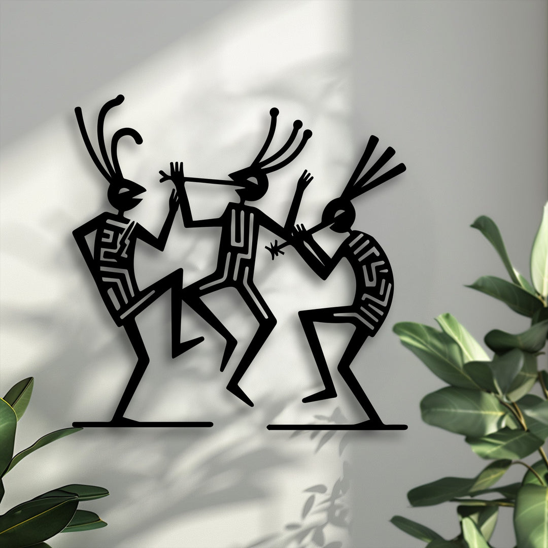 With low color saturation, this metal artwork features striking black tribal dance figures against a neutral wall, subtly enhanced by hints of vibrant green foliage.