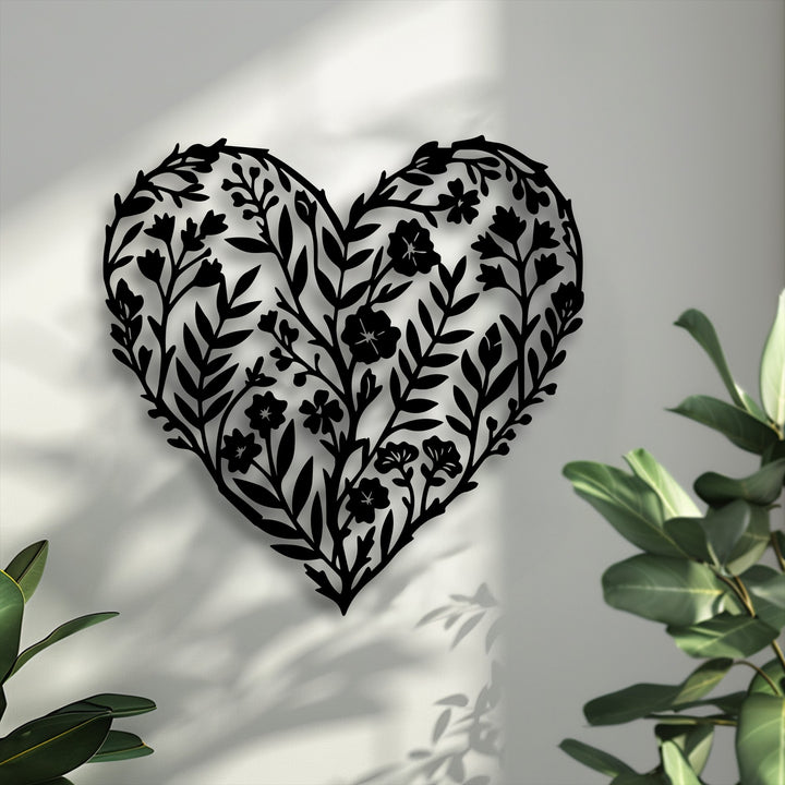 A vertically oriented, heart-shaped metal wall decor features an intricate botanical silhouette design on a plain white wall.