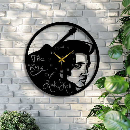 A black metal artwork wall clock, featuring Elvis Presley's guitar, creates layered depth on a white brick wall, framed by green plants.