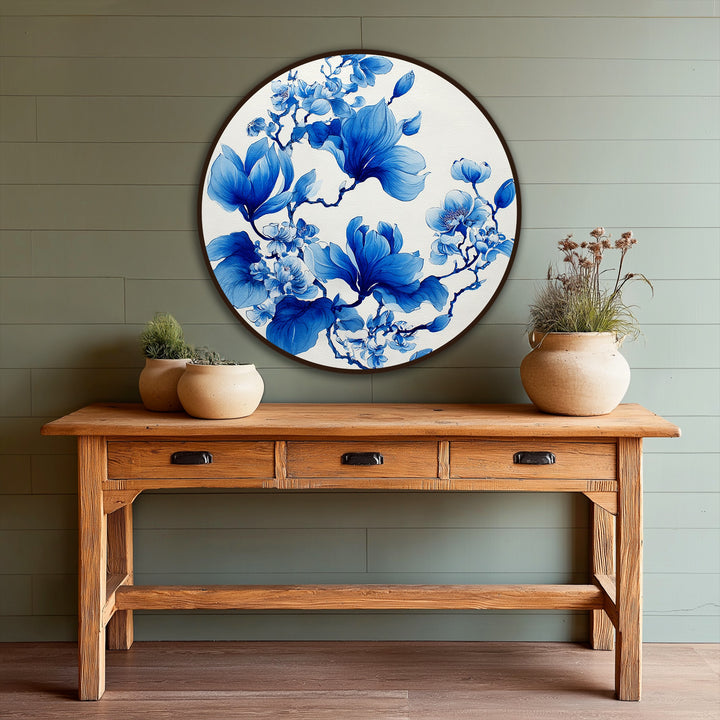 Set against a horizontally paneled muted green backdrop, an elegant round oil painting reproduction print featuring blue magnolia blossoms is displayed above a wooden console table.