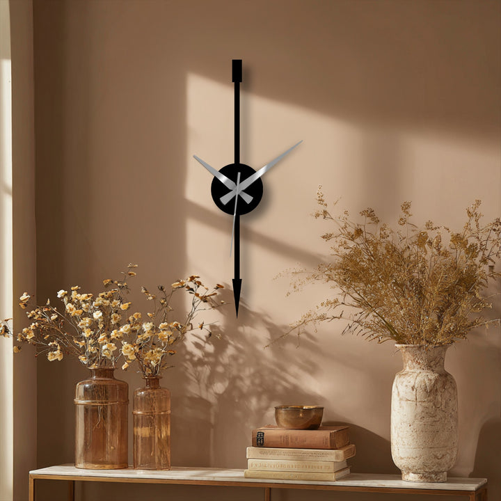 Illuminating the beige wall is the light, the softest element, highlighting the minimalist vertical arrow modern metal wall decor clock casting shadows beside elegant vases.