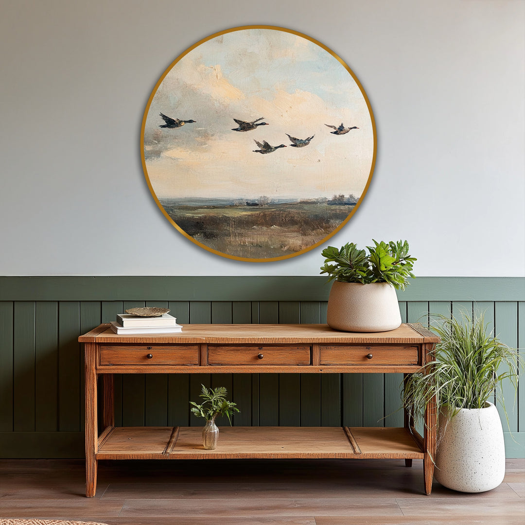 In the foreground, a rustic wooden console table displays a stack of books and greenery, complementing the round vintage oil painting home decor of flying ducks above.