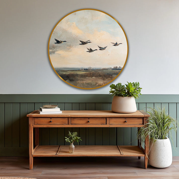 In the foreground, a rustic wooden console table displays a stack of books and greenery, complementing the round vintage oil painting home decor of flying ducks above.