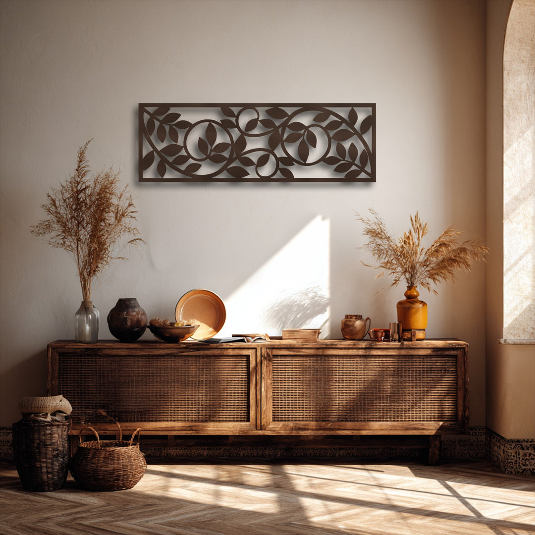 Sunlight brightly illuminates the wall behind a dark brown unique metal wall art panel featuring a flowing botanical vine pattern, creating stark shadows.