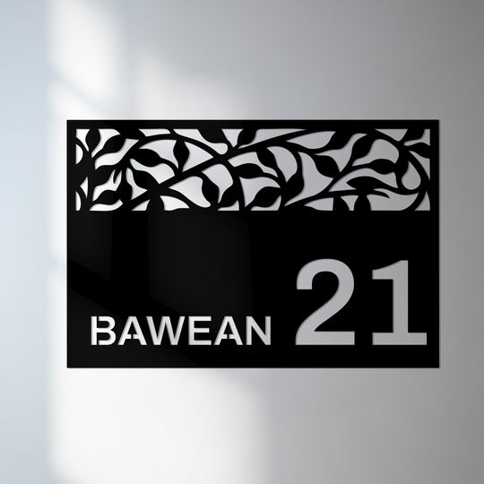The decorative metal art address sign shows a sharp transition from its leafy patterned top to its smooth, darker bottom, highlighting the contrast in colors.