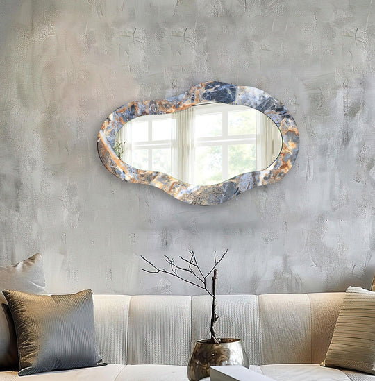 A textured gray and beige couch sits beneath a wavy, abstract wall mirror for wall decor with an intricate stone-like frame, reflecting a sunlit window.