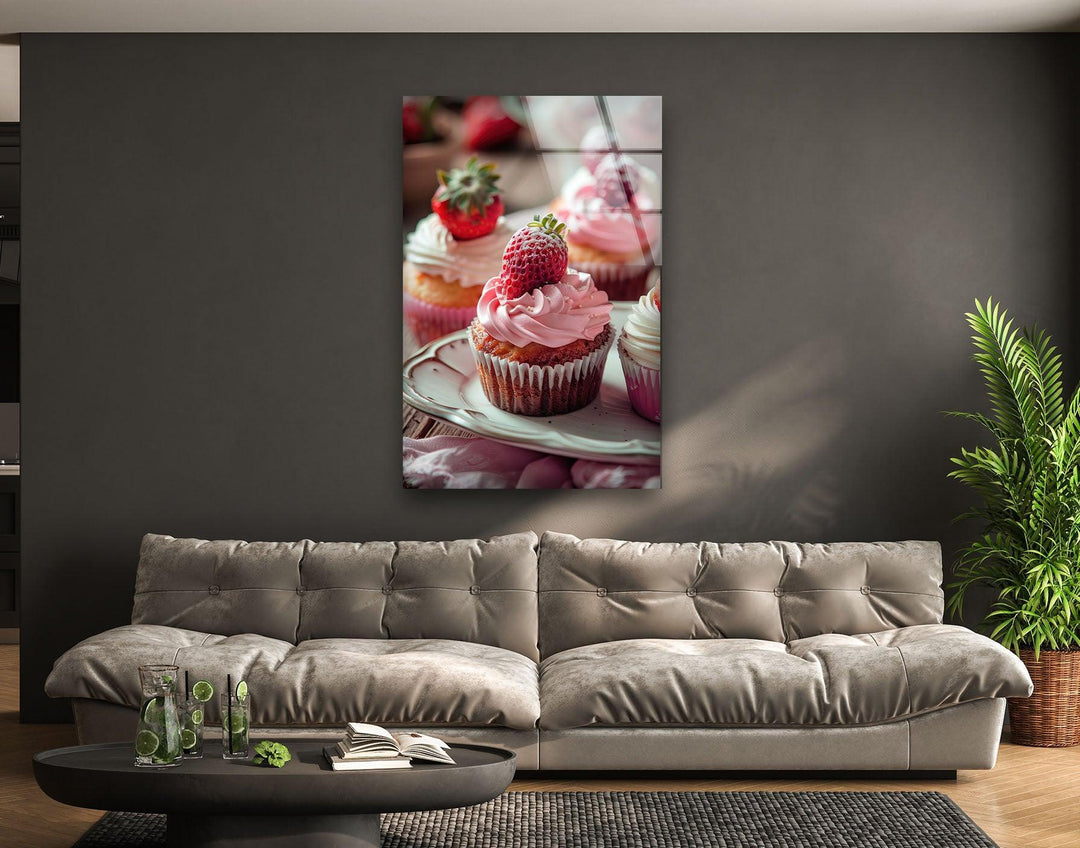 Vertical glass wall art featuring a close-up of pink frosted strawberry cupcakes displayed above a gray sofa in a modern living room.