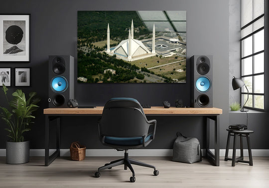 Foreground wood desk with a black chair and gaming peripherals highlights the framing of the hanging wall art featuring an aerial view of a mosque.