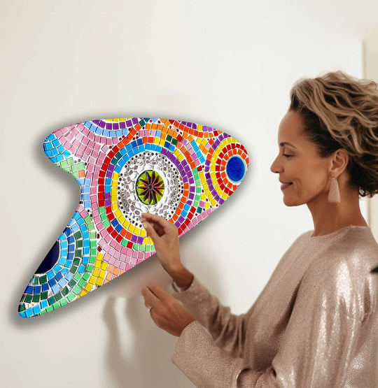 A woman admires a colorful, iridescent pink mosaic irregular glass wall art piece, the brightest element on a white wall.