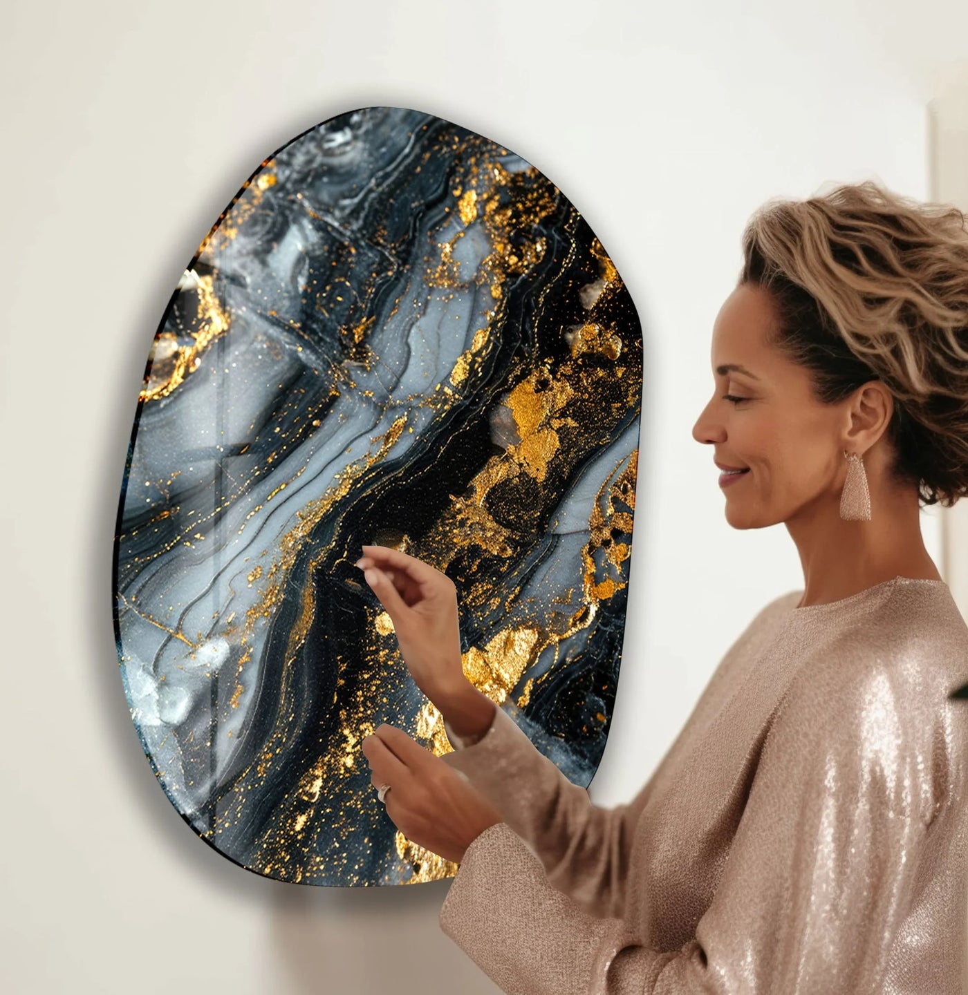 A woman admires the interplay of light and shadow across the swirling dark grey marble and gold leaf of this oval glass wall art, a striking addition to abstract wall prints.