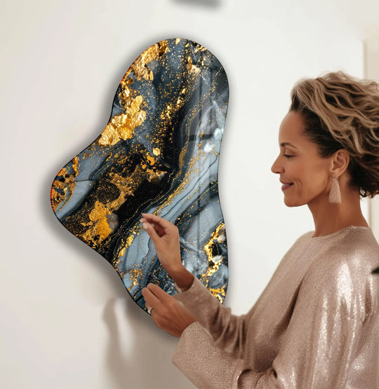 A woman admires a piece of asymmetric glass wall artwork dominated by deep grays and shimmering gold accents.