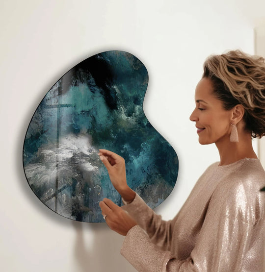 A woman's hand touches a contemporary abstract irregular decorative wall art, its swirling deep greens and grays dramatically highlighted by soft ambient light and subtle shadows.