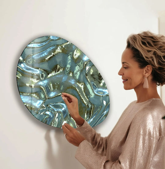 A woman in a sparkling top touches a modern green abstract oval glass wall art piece featuring rippling textures and metallic accents.
