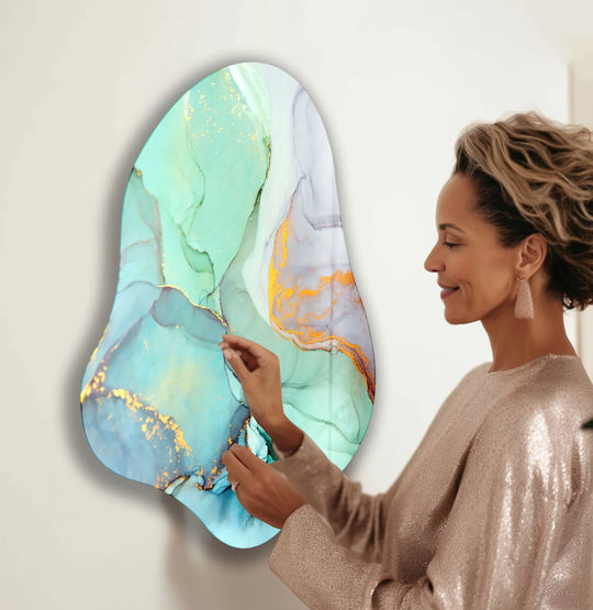 A woman with a sparkly shirt adjusts a modern, fluid-shaped piece of glass wall artwork with turquoise and gold marble-like details.
