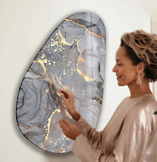A woman admires the lustrous, metallic gold and grey ink finish on an asymmetrical oval abstract glass hanging wall art.