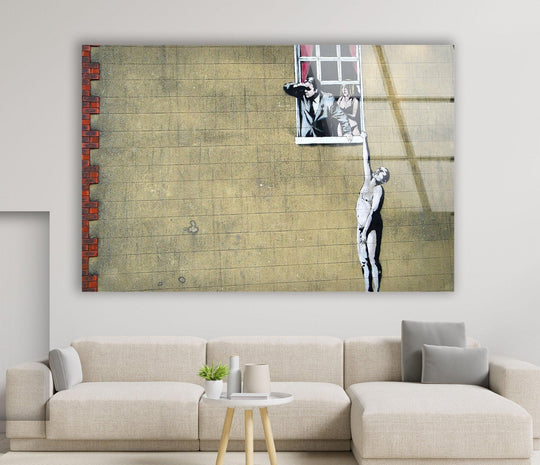 Banksy Well Hung Lover glass wall art is a pop art piece with the naked man mural on a yellow wall and displayed over a modern sofa