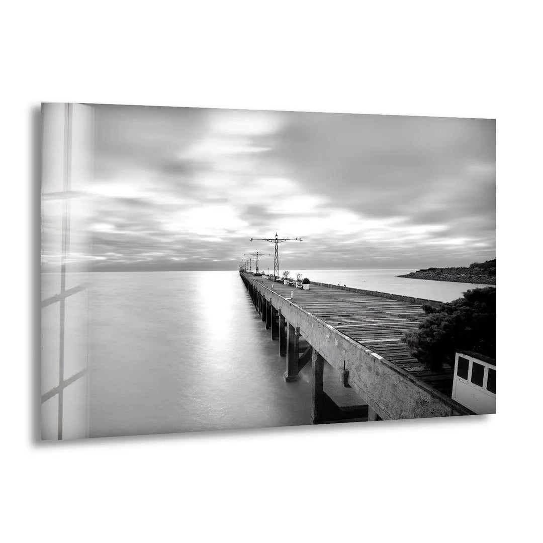 Long wooden pier extending into calm water under soft blurred clouds in black and white glass wall art.