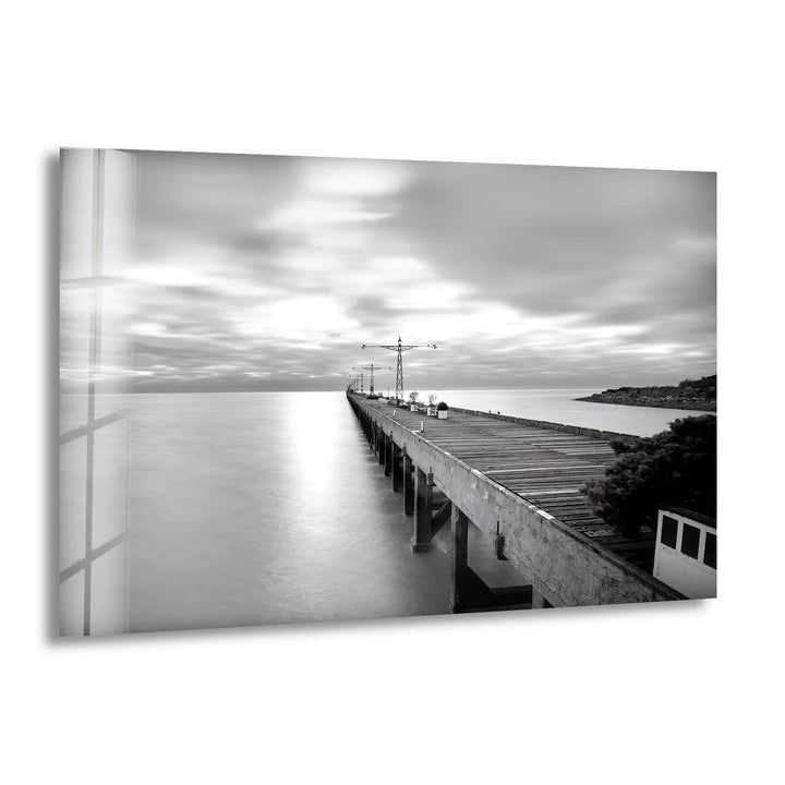 Long wooden pier extending into calm water under soft blurred clouds in black and white glass wall art.
