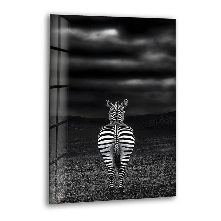 Black and white glass wall art featuring a zebra standing alone, viewed from behind under a dramatic cloudy sky.
