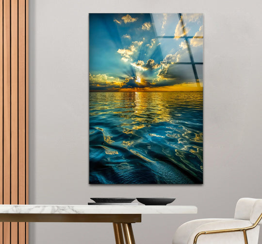 Glass wall art featuring a vertical ocean sunset with layered golden sky and deep blue water reflections.