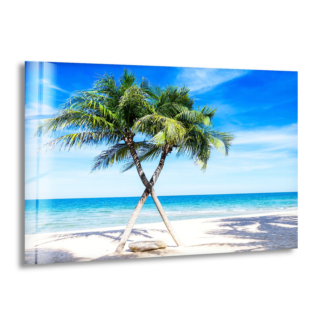 Tropical beach scene with two crossed palm trees under a bright blue sky, printed on glass wall art.
