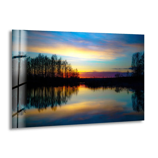 A striking glass print showcases the foreground detail of a serene lake reflecting a vivid sunset and silhouetted trees in tranquil home decor.