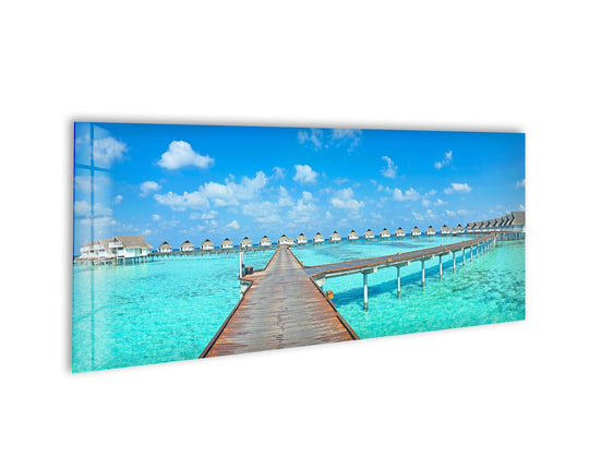 Tropical Maldives glass wall art showcases a wooden pier and turquoise water and features a serene ocean view and panoramic landscape above a white wall