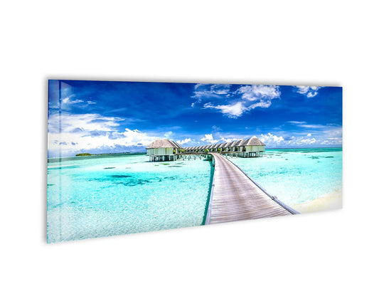 Maldives Beaches Landscape glass wall art showcases a wooden walkway and overwater bungalows and vibrant turquoise water and a blue sky and white clouds