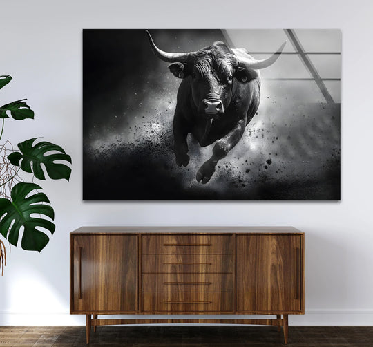 Dramatic light and shadow highlight a powerful bull charging through dust in this black and white glass wall art, a striking addition to any space.