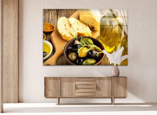Horizontal glass wall art depicting olives and bread arranged on a wooden board above a light wood console in a modern living room setting.
