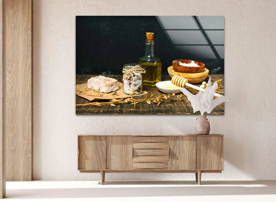 Horizontal glass wall art depicting a still life with olive oil, honey, and a white dessert on a rustic wooden surface above a sideboard.