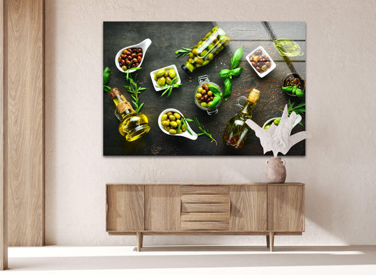 Horizontal glass wall art depicting a still life of olives, oil bottles, and basil on a dark surface, displayed above a wooden sideboard in a modern living room.
