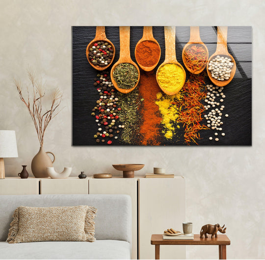 Horizontal glass wall art featuring wooden spoons filled with colorful spices arranged on a dark slate background above a neutral sofa in a living room.